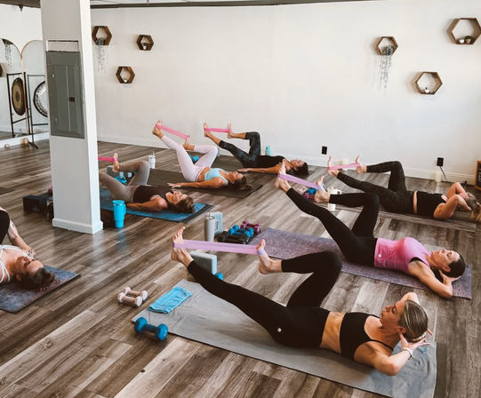 Group pilates class in a bright fitness studio: women on mats doing leg raises with pink resistance bands, dumbbells, towels and water bottles on a wood floor.