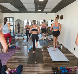 Group fitness class in a mirrored studio with women in activewear balancing on one leg on mats, dumbbells and yoga props on the floor.