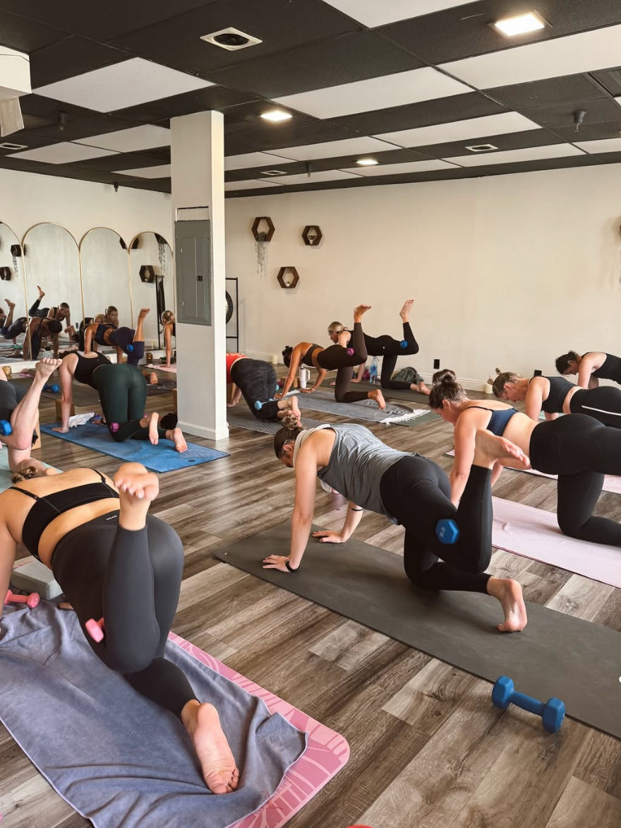 Group mat workout in a bright fitness studio — participants doing glute-kickback exercises with small dumbbells/ankle weights, mirrored wall and wood-look floor.