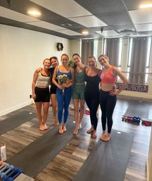 Six women smiling barefoot on yoga mats in a bright indoor yoga studio during a small group yoga/fitness class, one holding a bouquet, wearing workout clothes with dumbbells and mats visible.