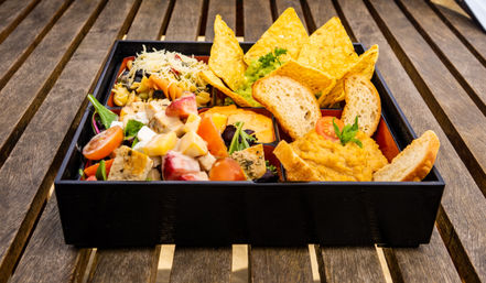 Black bento-style tray of colorful appetizers on a wooden outdoor table: mixed greens with cherry tomatoes and diced protein, cheesy pasta salad, guacamole with tortilla chips, toasted baguette slices and a dollop of hummus garnished with herbs.