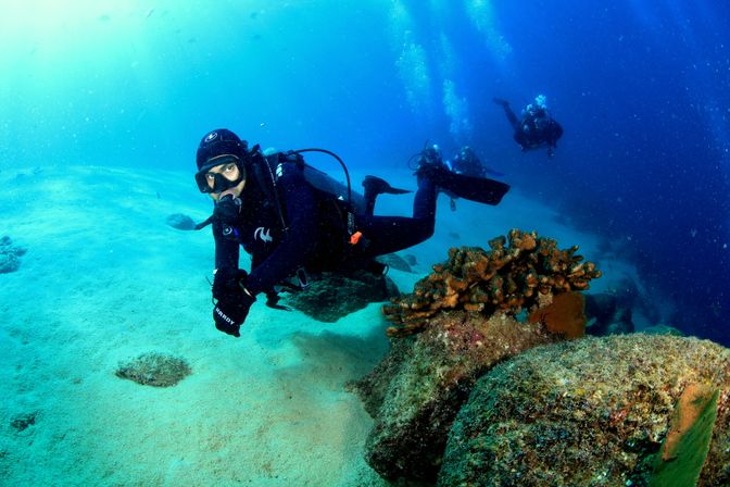 Scuba diver in black wetsuit hovering above sandy seabed beside coral-covered rocks, with two other divers and rising bubbles in the clear blue ocean — underwater scuba diving coral reef scene