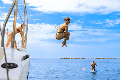 Person mid-cannonball jumping off a white boat into clear blue ocean, another person leaning on the boat railing and a paddleboarder nearby under a sunny sky