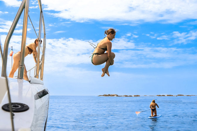 Person mid-cannonball jumping off a white boat into clear blue ocean, another person leaning on the boat railing and a paddleboarder nearby under a sunny sky