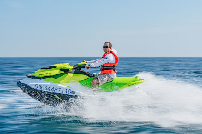 Rider in an orange life vest and sunglasses speeding on a bright green jet ski across blue open water, white spray trailing behind on a sunny day.