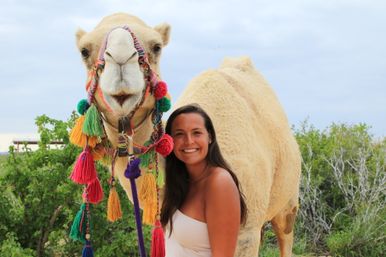 Smiling woman posing beside a cream-colored camel wearing a colorful tassel bridle in a scrubby desert landscape.
