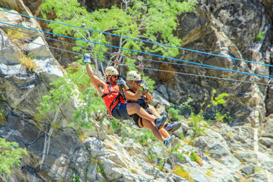 Tandem zipline riders in helmets and harnesses soaring above a rocky canyon with green trees — outdoor mountain zipline adventure
