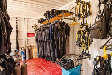 Dive shop storage with rental scuba gear: black wetsuits hanging on a rack, neoprene boots on a shelf, scuba tanks on the red floor and yellow-hosed regulators neatly hung on a white container wall — ready for the next dive.