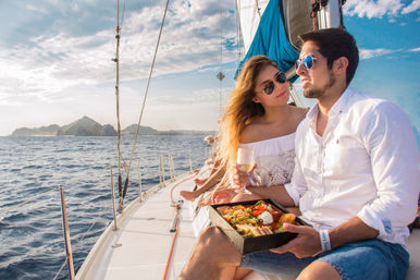 Couple in sunglasses enjoying a picnic and wine on a sailboat cruising the ocean near rocky coastal islands under a blue sky