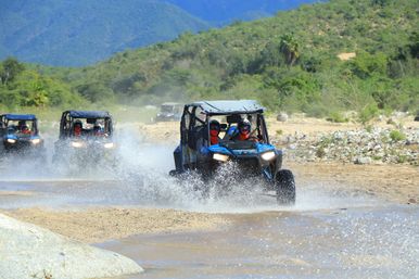 Group of off-road UTVs splashing through a shallow desert river on an outdoor adventure, riders in helmets, sandy riverbed with green foothills and blue mountain backdrop