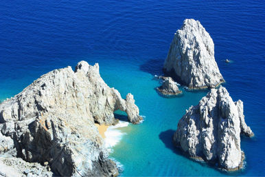 Aerial view of sunlit coastal rock formations and a natural sea arch rising from turquoise to deep-blue ocean with a tiny sandy cove