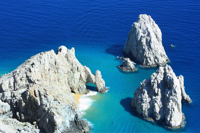 Aerial view of sunlit coastal rock formations and a natural sea arch rising from turquoise to deep-blue ocean with a tiny sandy cove