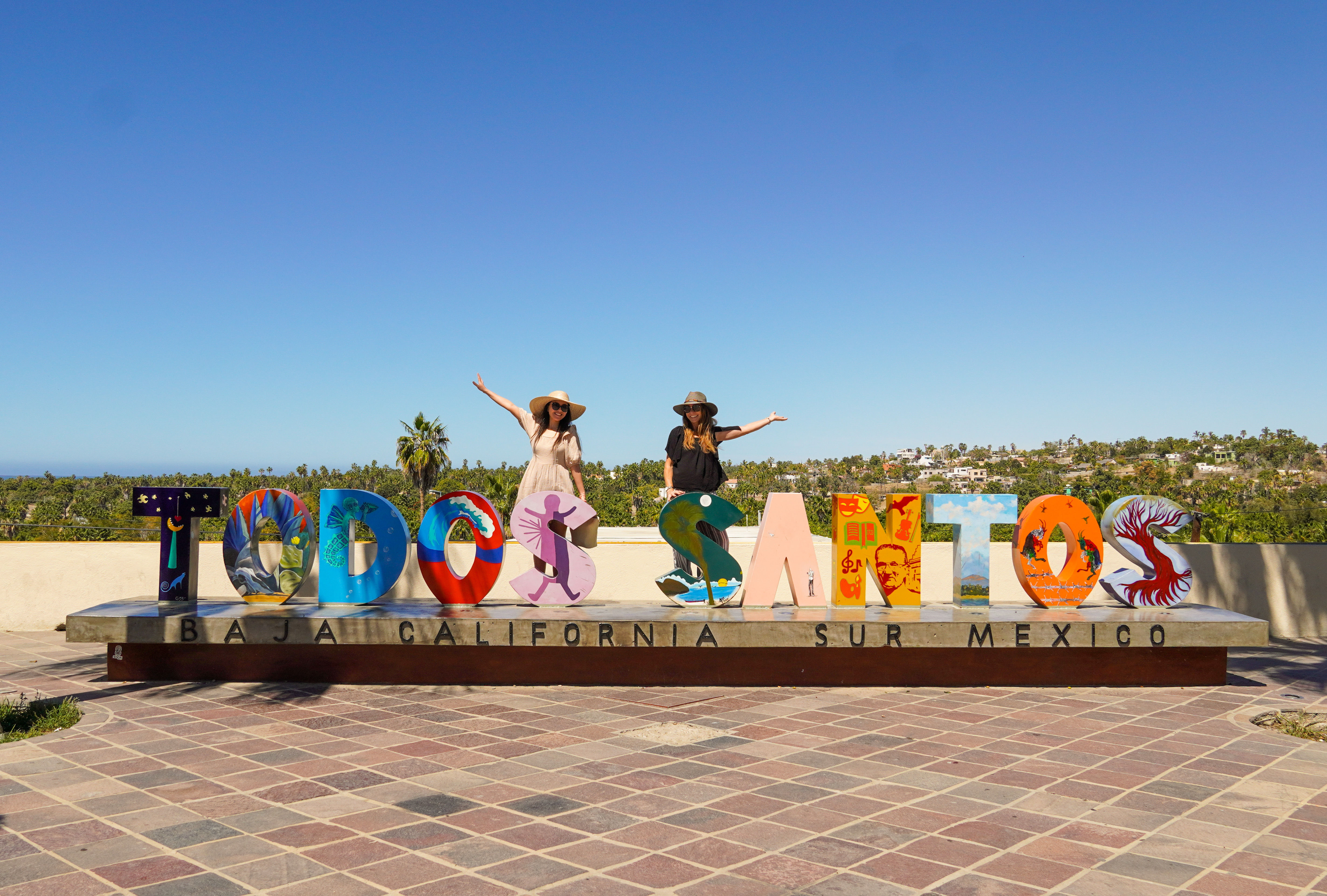 Colorful Todos Santos landmark sign with painted letters on a plaza, two people posing on the letters under a bright blue sky with palm trees and coastal town in the background in Baja California Sur, Mexico.