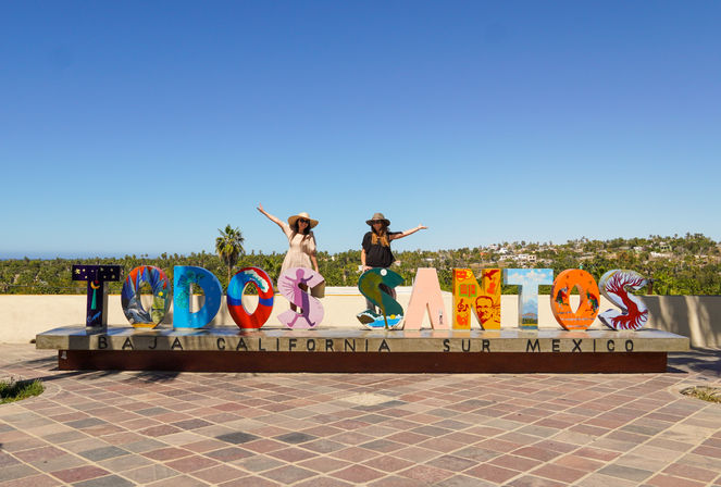 Colorful Todos Santos landmark sign with painted letters on a plaza, two people posing on the letters under a bright blue sky with palm trees and coastal town in the background in Baja California Sur, Mexico.