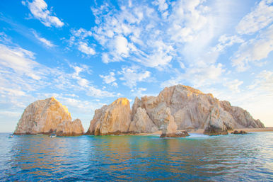 Sunlit coastal sea stacks and natural rock arch rising from turquoise ocean at golden hour under a bright blue sky with scattered clouds