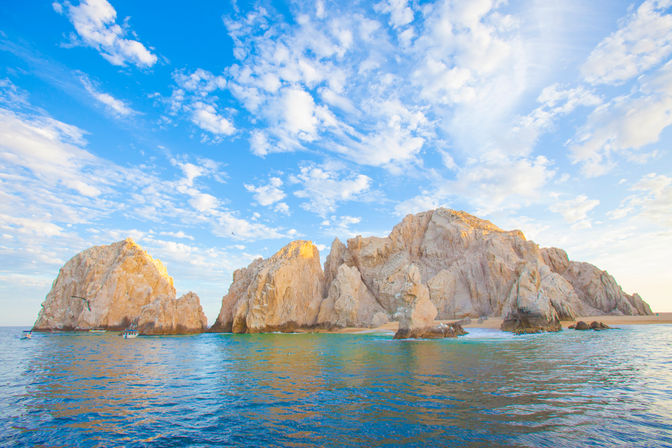 Sunlit coastal sea stacks and natural rock arch rising from turquoise ocean at golden hour under a bright blue sky with scattered clouds