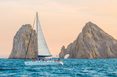 White sailboat gliding past a dramatic sea arch and rugged rocky islets at sunset over turquoise ocean, passengers enjoying a coastal cruise.