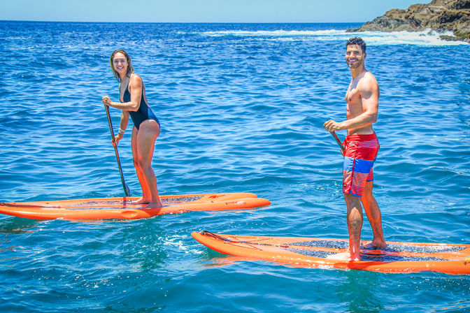 Two smiling adults stand on bright orange stand-up paddleboards in clear blue coastal waters near a rocky shoreline on a sunny day.