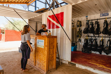 Woman in wetsuit chatting with staff at a beachside scuba dive shop check-in inside a shipping container, red diver flag and hanging scuba gear visible.