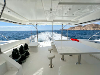 Aft deck of a white yacht cruising on deep-blue ocean near a rocky coastline — shaded table and stools, bench seating, three black sea scooters stored left, red life jacket and wake trailing behind.