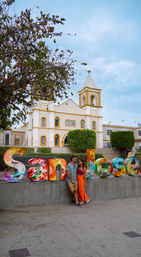 Couple posing by vibrant painted 'San José' letters in a plaza, with a yellow-and-white colonial church, trimmed topiary, and a cloudy blue sky overhead.