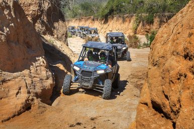 Line of off-road UTV side-by-side buggies navigating a narrow sandy desert canyon with orange rock walls, riders in helmets and bandanas on a rugged adventure tour