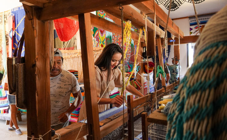 Woman weaving a blue patterned textile on a large wooden handloom in a vibrant artisan workshop surrounded by colorful woven rugs and craft supplies.