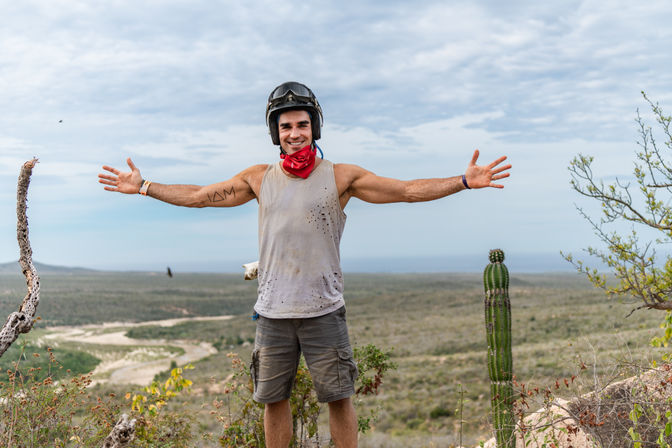 Off-road rider wearing a helmet and red bandana stands arms wide on a desert hill beside a tall cactus, overlooking a winding dirt trail and distant ocean.