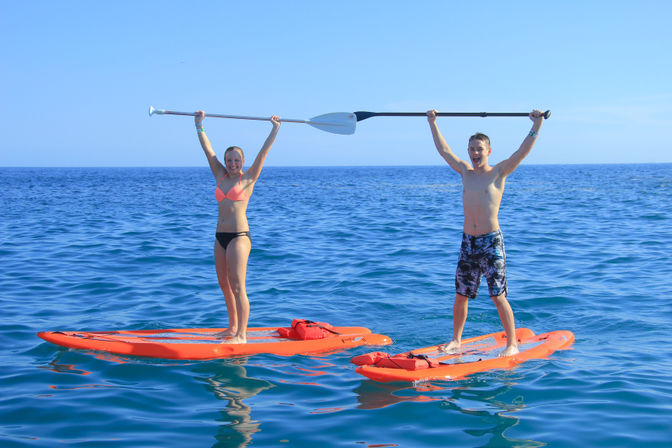 Two people standing on bright orange stand-up paddleboards in a calm blue ocean, holding a shared paddle overhead and smiling on a sunny day.