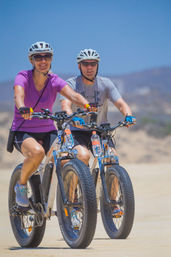 Two smiling cyclists in helmets and sunglasses riding fat-tire bicycles along a sunny sandy beach trail under a clear blue sky.