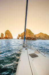 Sailboat bow gliding toward a sunlit coastal rock arch and rugged sea stacks at golden sunset over deep blue water