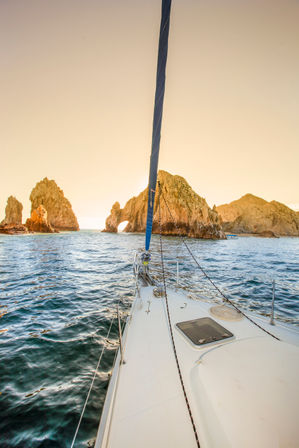 Sailboat bow gliding toward a sunlit coastal rock arch and rugged sea stacks at golden sunset over deep blue water