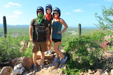 Three people in helmets and colorful bandanas pose on a rocky coastal desert overlook surrounded by cacti, green scrub and pink wildflowers with a distant ocean and blue sky.