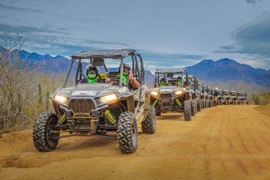 Line of off-road side-by-side UTVs on a dusty desert trail with riders in helmets and green bandanas and rugged mountains in the background