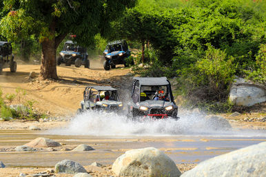 Action-packed group of off-road UTV side-by-sides splashing through a shallow river on a dusty trail, drivers in helmets, green brush and boulders nearby.