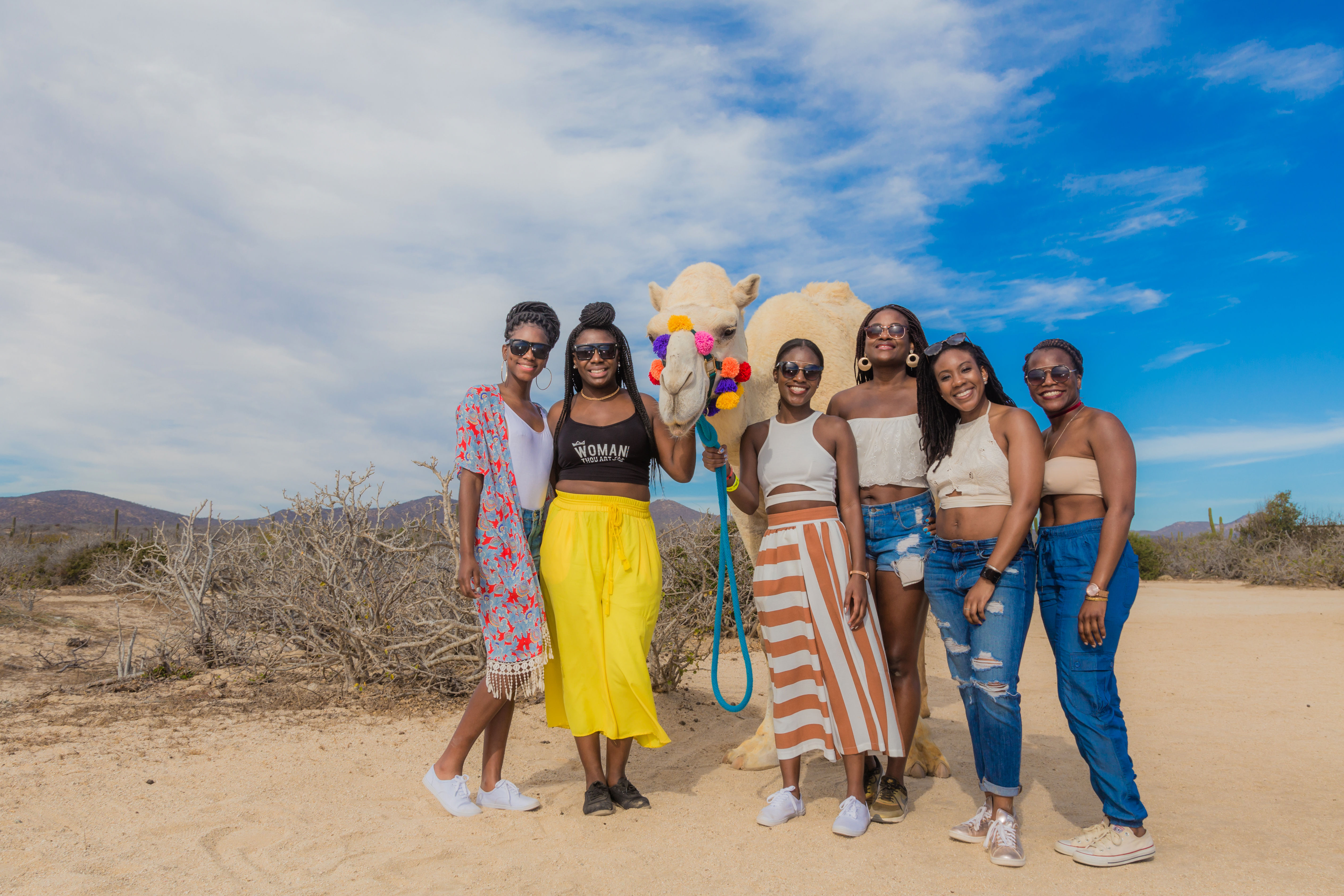 Six women posing with a colorful pom-pom–decorated camel in a sunny desert landscape under a bright blue sky — playful group vacation photo in casual summer outfits.