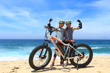 Two helmeted cyclists flexing their arms while posing with a fat-tire e-bike on a sunny sandy beach with blue ocean waves and a clear sky.