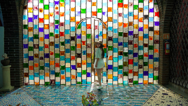 Woman in a straw hat and white dress stands in an arched opening of a vibrant stained-glass wall, colorful light reflecting across patterned mosaic tiles.