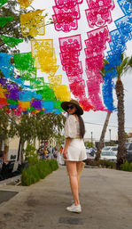 Colorful papel picado banners strung over a palm-lined street festival, a woman in a wide sun hat, sunglasses and white shorts strolling beneath.