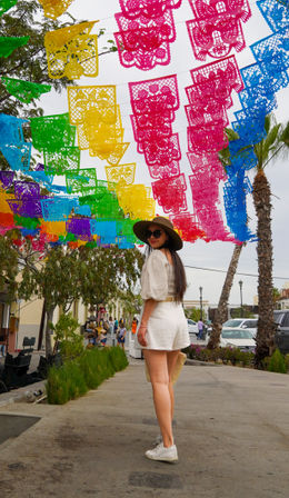 Colorful papel picado banners strung over a palm-lined street festival, a woman in a wide sun hat, sunglasses and white shorts strolling beneath.