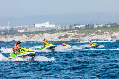 Four riders on neon-yellow jet skis in orange life vests skimming choppy blue sea near a rocky coastline with low coastal buildings in the background
