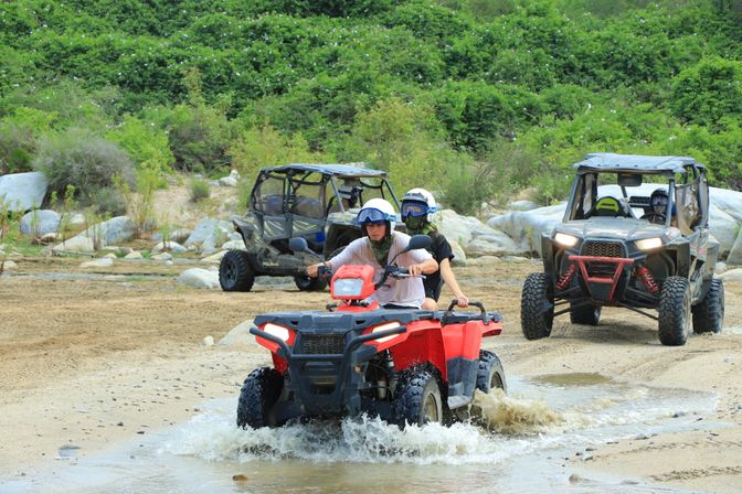 Two helmeted riders on a red ATV splashing through a shallow creek on an off-road trail, with two UTV side-by-sides, rocky banks and green hillside in the background — outdoor off-road adventure.