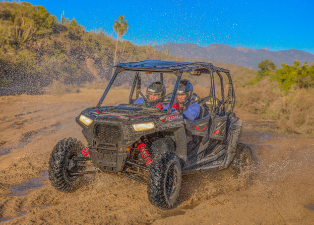Two helmeted riders in a four-seat off-road UTV kicking up mud on a sunny desert trail with palm trees and mountains in the background