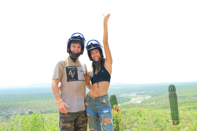 Two adventurers in off-road helmets pose at a desert overlook with tall cacti and a winding river in the green valley below; woman in a black crop top raises her arm in celebration.