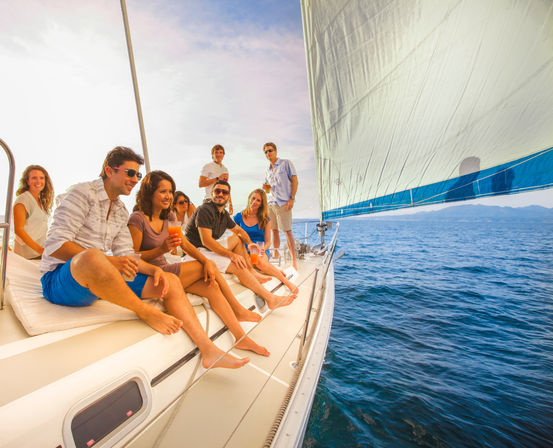 Group of friends on a sailboat with a large blue sail, enjoying drinks on sunny ocean waters