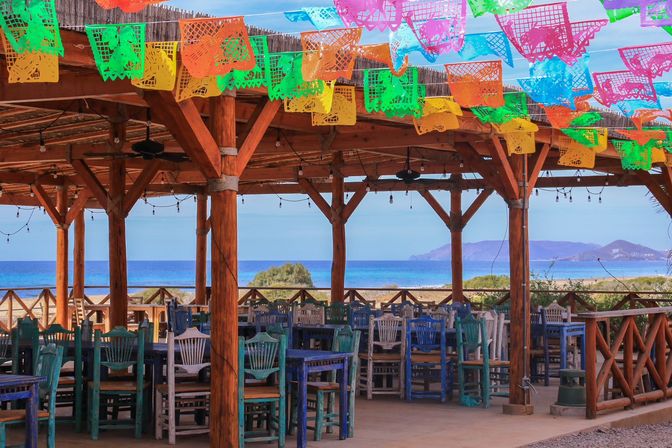 Beachfront outdoor dining under a rustic wooden pergola with vibrant papel picado banners, mismatched painted chairs and tables, and a clear turquoise ocean with distant islands on the horizon.