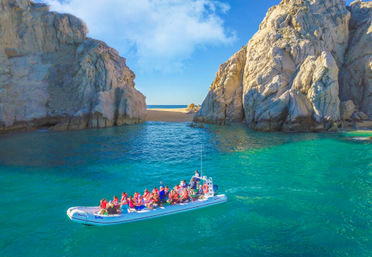 Inflatable tour boat with passengers in red life jackets gliding across turquoise water through a rocky coastal cove toward a sandy beach and open sea under a bright blue sky