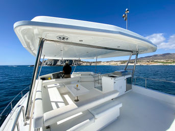 Sunlit yacht flybridge with white lounge seating, table with a drink, built-in grill and a person at the helm cruising blue coastal waters toward a rocky shoreline.