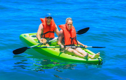Two people wearing orange life jackets paddling a bright green tandem kayak on vivid blue ocean water, smiling and enjoying a sunny, sunny-day paddle.