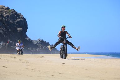 Cyclist in helmet and pads rides a fat-tire bike on a sandy beach near rocky cliffs, legs kicked out in a playful split while another rider follows along the ocean shoreline under a clear blue sky.